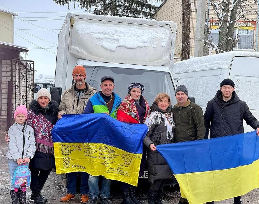 People holding two Ukrainian flags, one signed, in front of a snow-covered white truck.