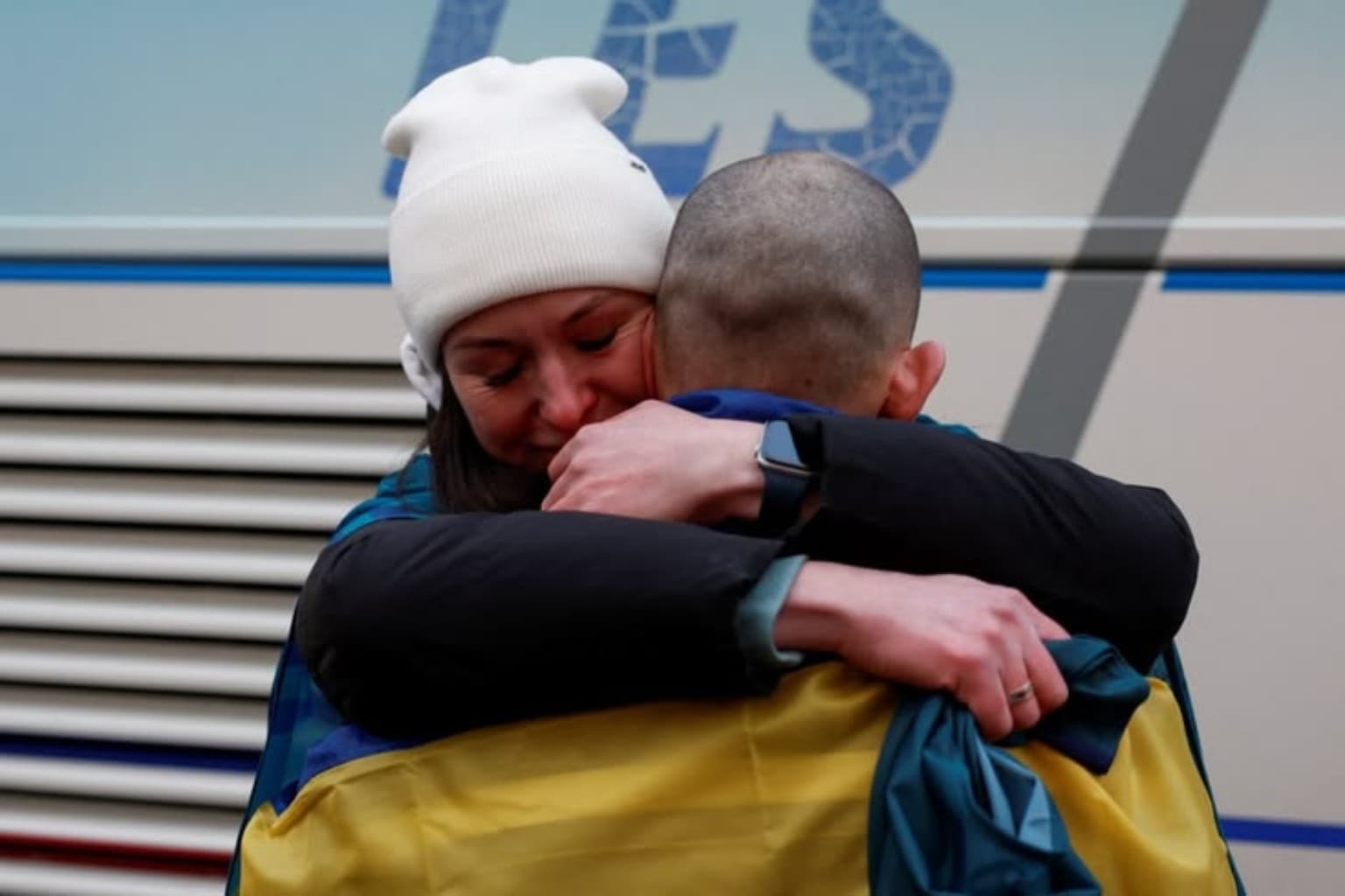 Woman in white beanie emotionally hugs a man draped in a Ukrainian flag.