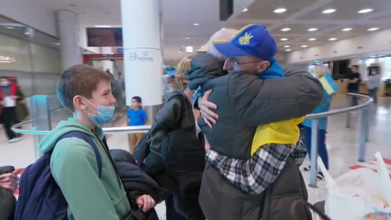 Man in a Ukrainian cap hugs a relative at an airport while a boy watches.