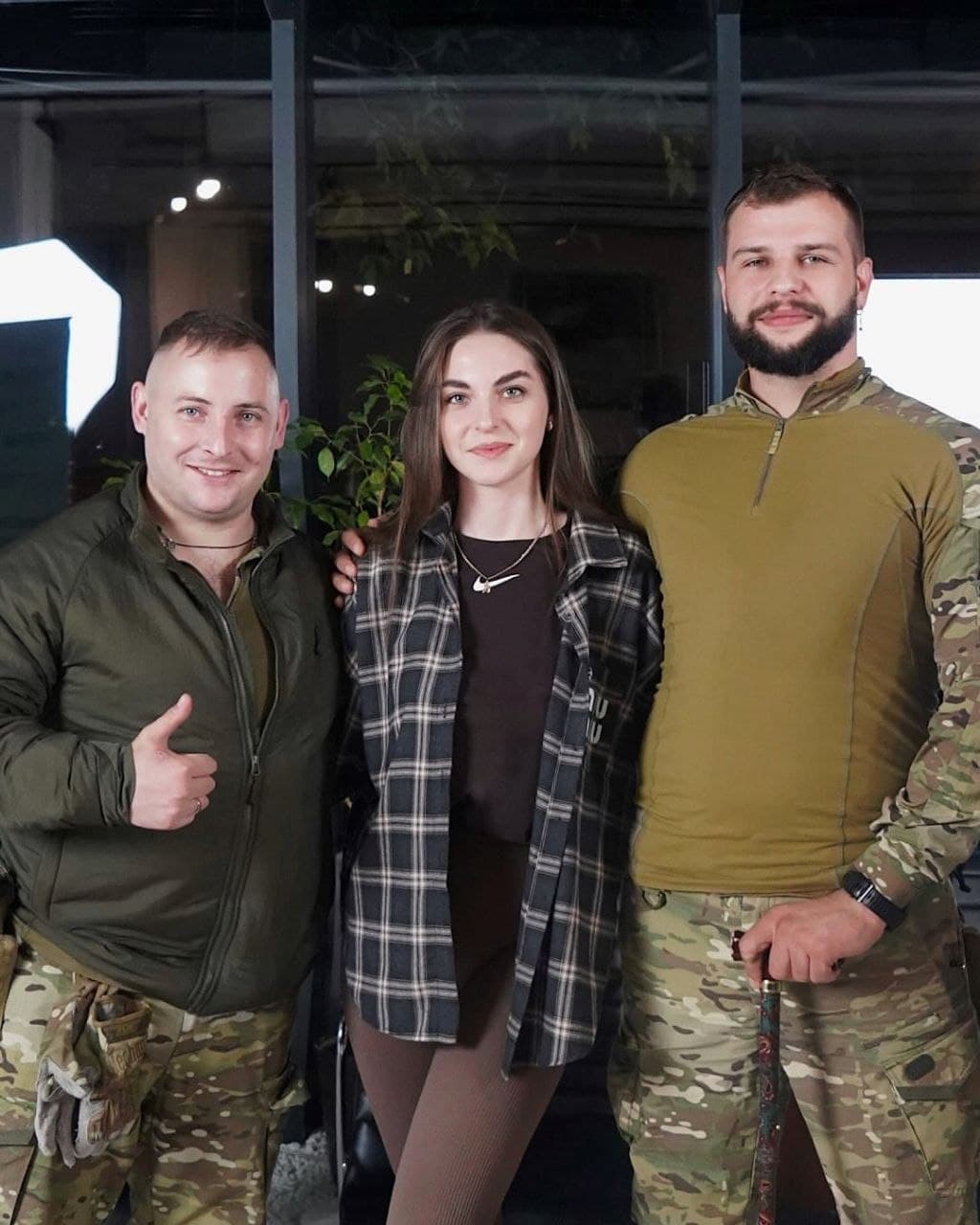 Woman in plaid shirt stands between two men in military uniforms, one giving thumbs up.